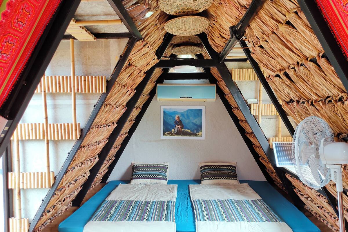 Habitación triangular con cama, almohadas, un ventilador y un cuadro en la pared.