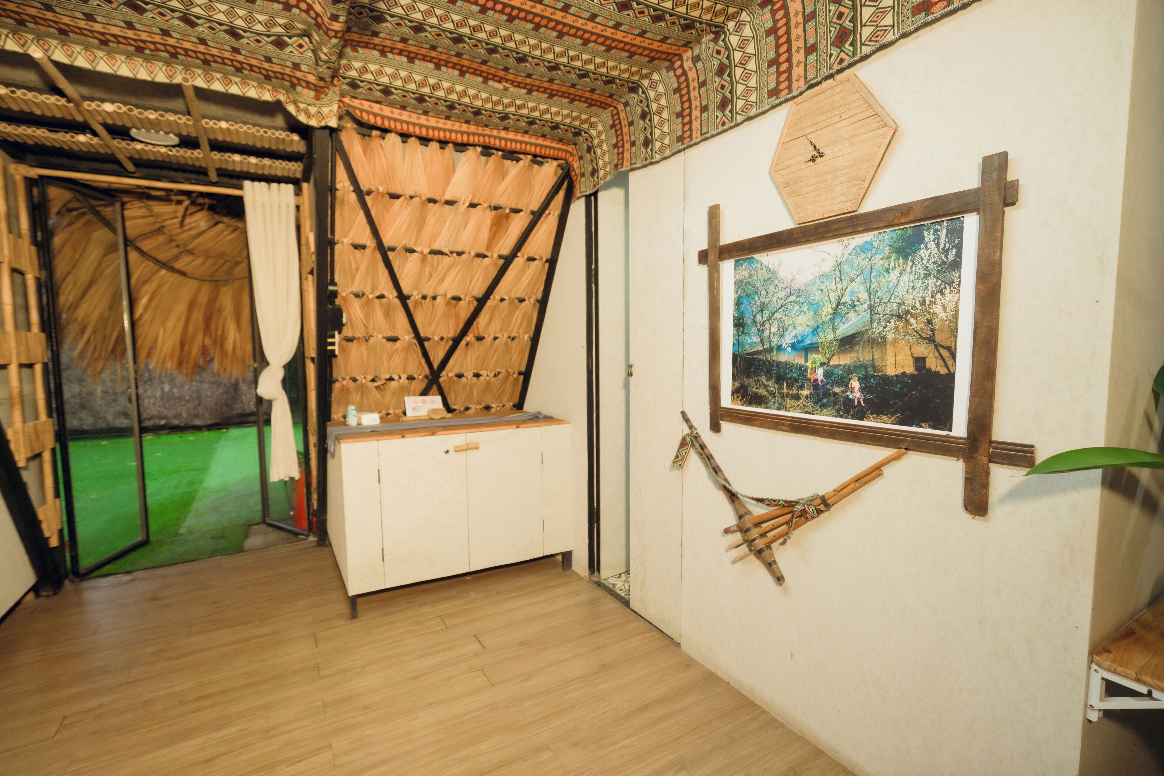 A room with a wooden floor, a thatched-roof entrance, a wooden cabinet, a framed picture on the wall, and a hanging hammock.