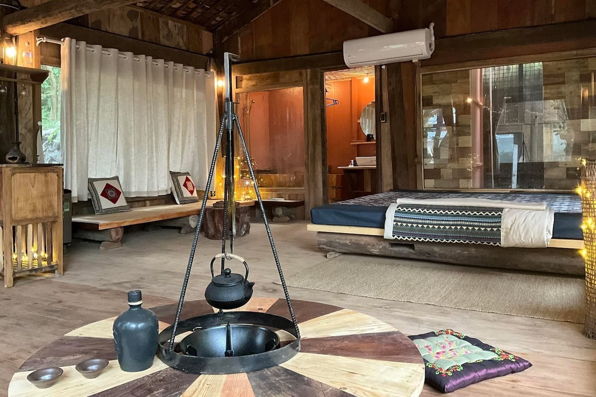 Una habitación rústica con una cama, un hervidor colgante, una mesita con cuencos y un ventanal grande.