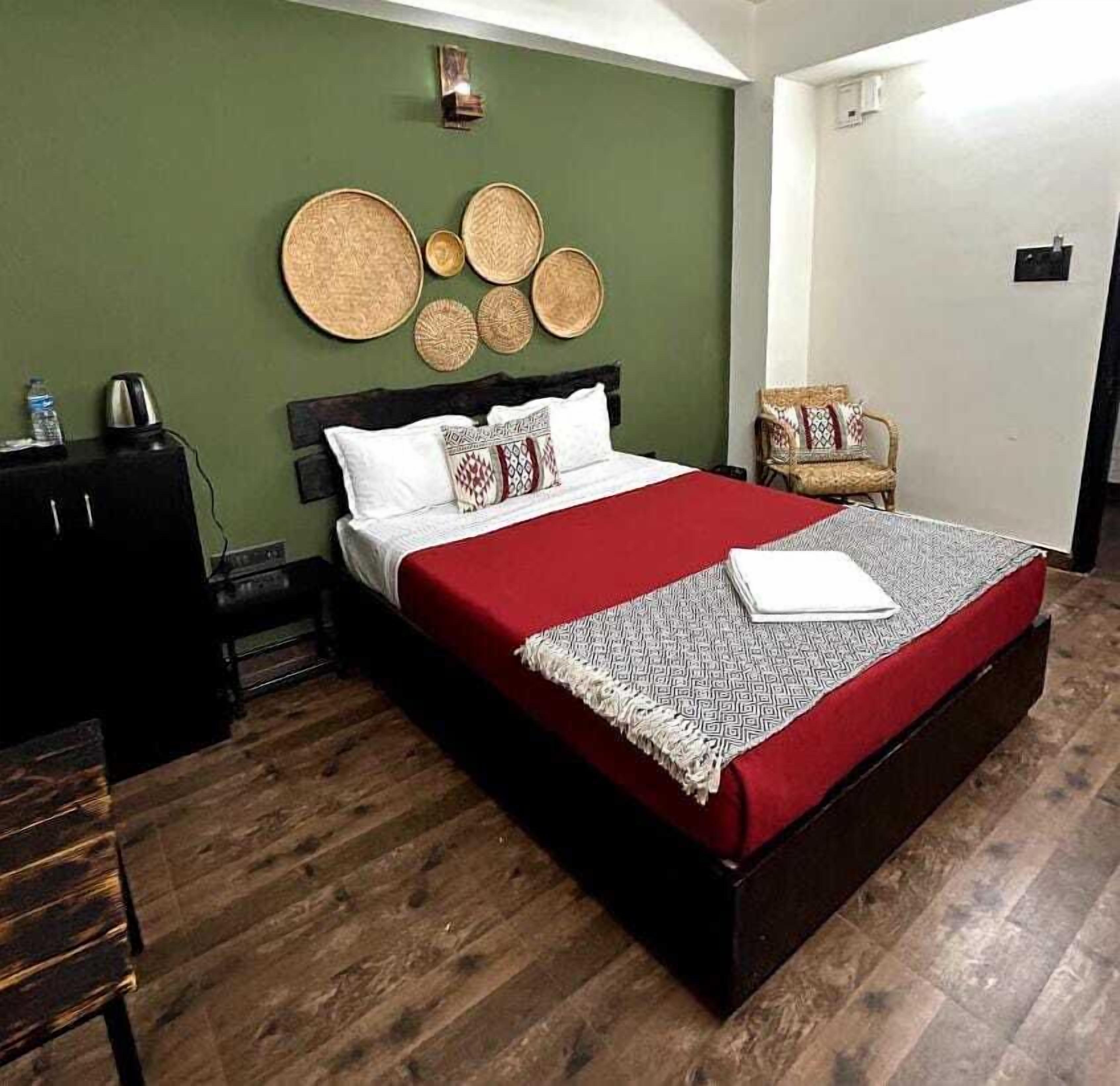 Una cama bien hecha con una colcha roja y un plaid estampado, una mesita de noche de madera con una tetera, una lámpara de pared y una silla con asiento de mimbre.