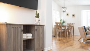Un meuble en bois sur lequel est fixé un téléviseur à écran plat, une plante en pot posée sur le dessus, et un coin repas avec des chaises et une table en bois à l’arrière-plan.