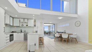 A modern kitchen with white cabinetry, a central island, and a dining area with a round table and chairs. - The Lookout Retreat - Woorim (City of Moreton Bay)