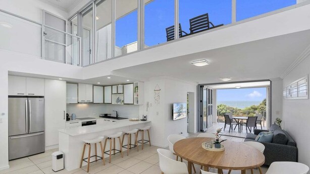 A modern kitchen with a central island, a dining area with a round table, and a view of the outdoors. - The Lookout Retreat - Woorim (City of Moreton Bay)