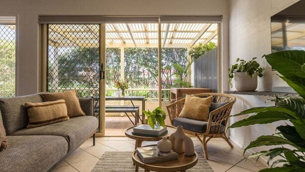 A modern living room with a sofa, armchair, and coffee table, a sliding glass door leading to an outdoor patio with plants and a table. - Carmine - Fiesta Palms (Byron Bay)