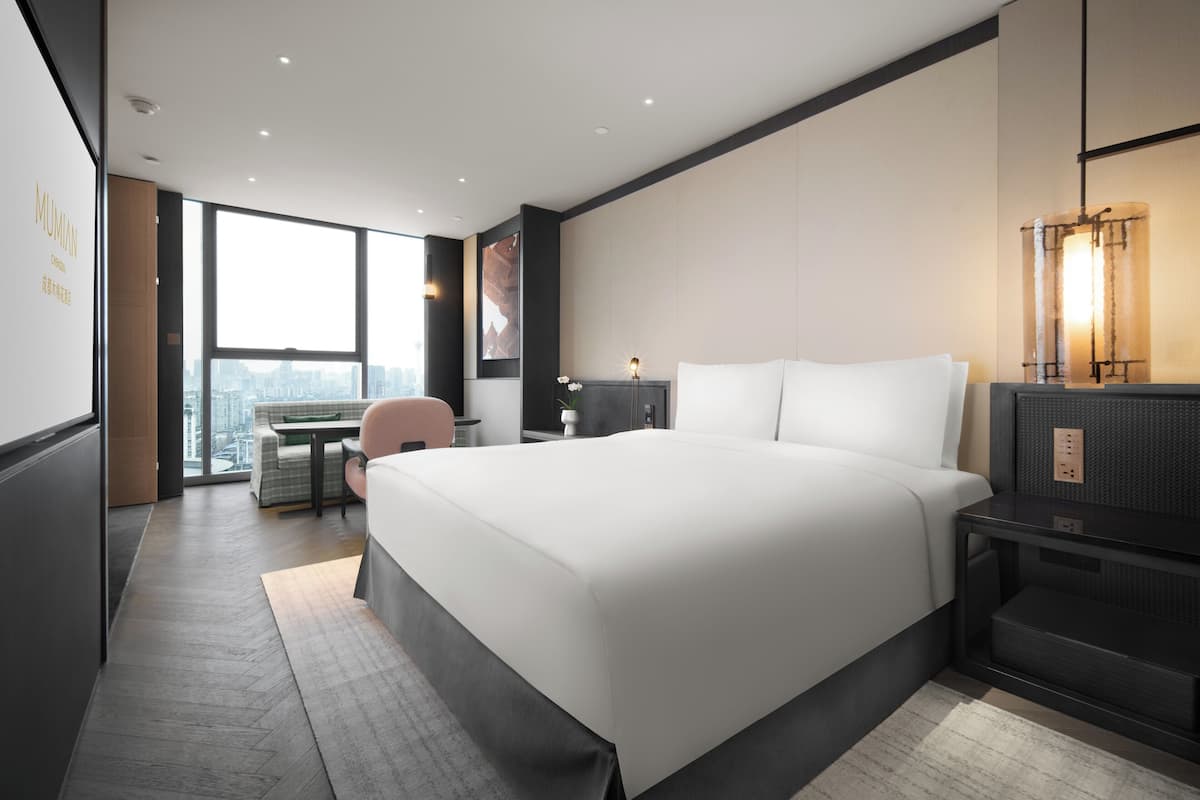 A modern hotel room with a large bed, a desk, a chair, and a city view.