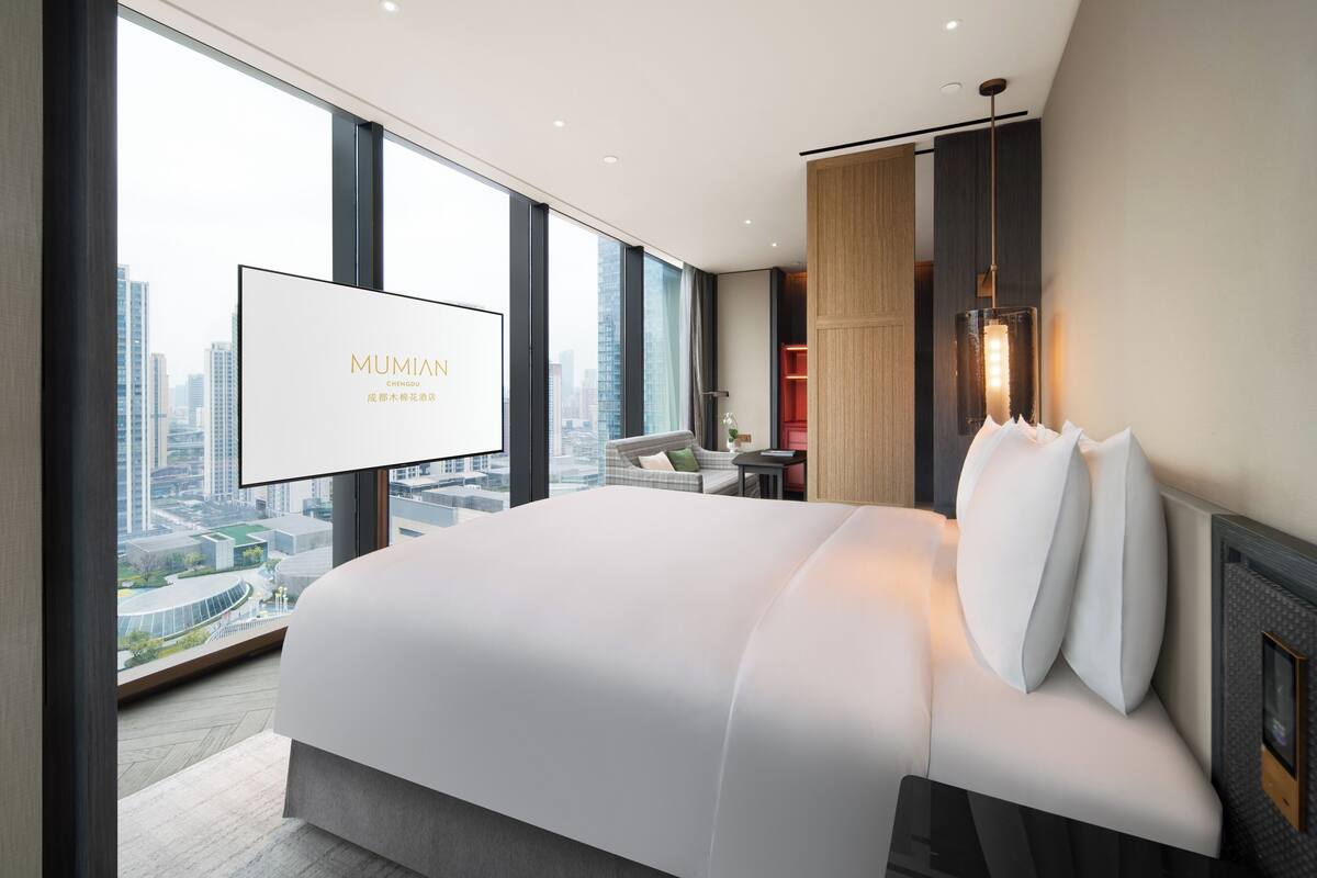 A modern hotel room with a large bed, a city view, a mounted TV, and a unique wall-mounted light fixture.