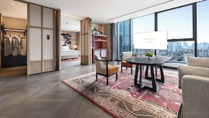 A modern hotel room with a large city view, a dining area with a table and chairs, and a wardrobe with hanging robes.