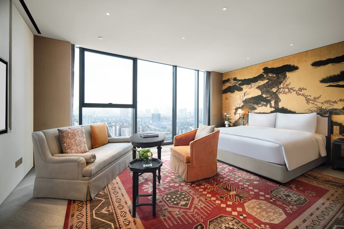 A modern hotel room with a large bed, a sofa, a small table with a vase, and a large window offering a city view.