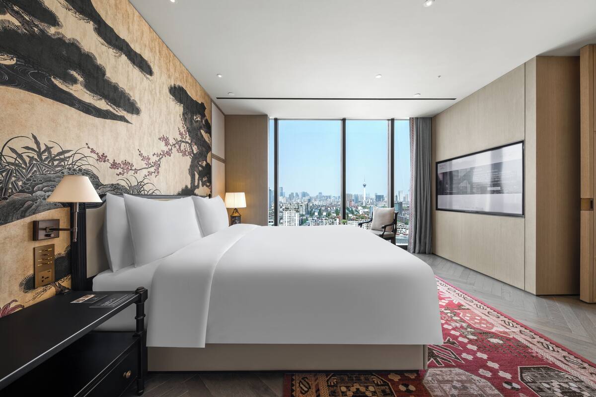 A modern hotel room with a large bed, a city view, a mounted artwork, and a television.