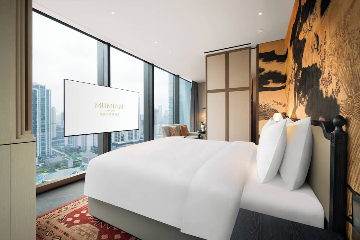 A modern hotel room with a large bed, a city view, and a large screen displaying the hotel name.