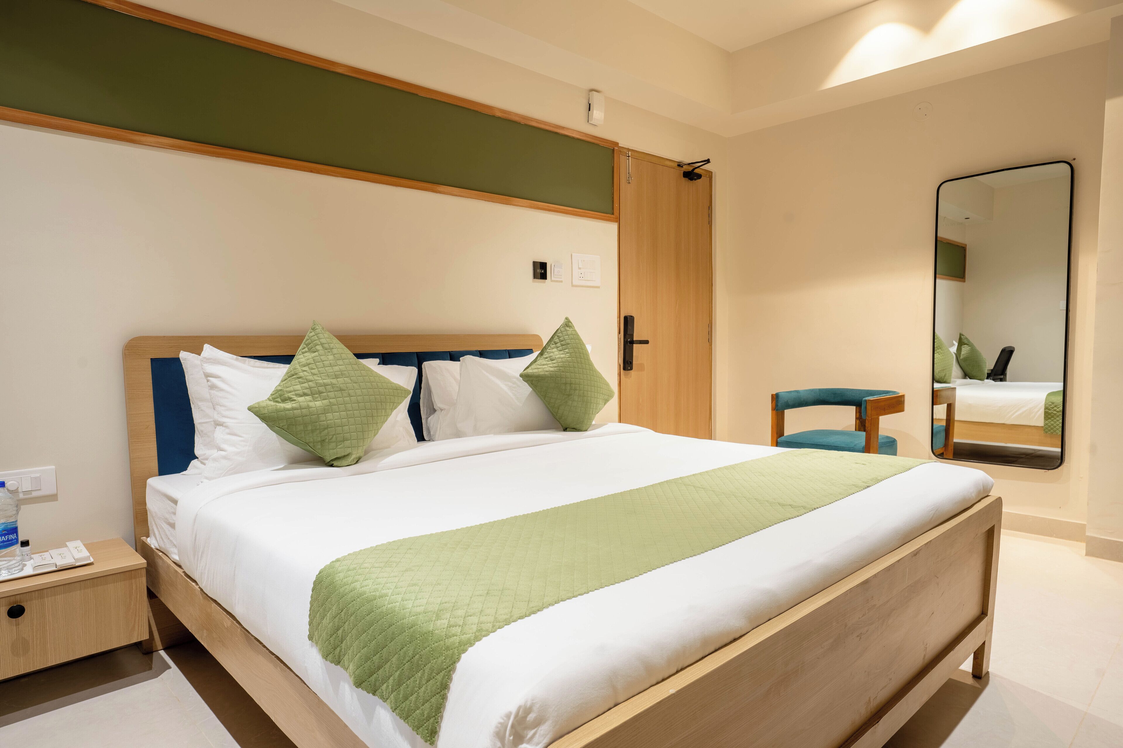 A neatly made bed with a green blanket, a wooden headboard, and two pillows. There is a wooden nightstand with a water bottle and a remote control. A mirror is on the wall, and a wooden door is visible.