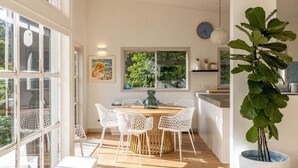 Una cocina luminosa con una mesa redonda de madera y sillas blancas, ventanas amplias y una planta en maceta.