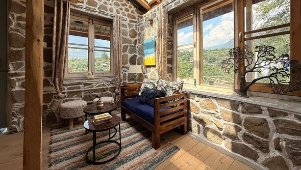 A cozy room with a stone wall, wooden beams, a sofa, a small table, and a view of the outdoors. - KALA STUDIO AND STAY (Kullu)