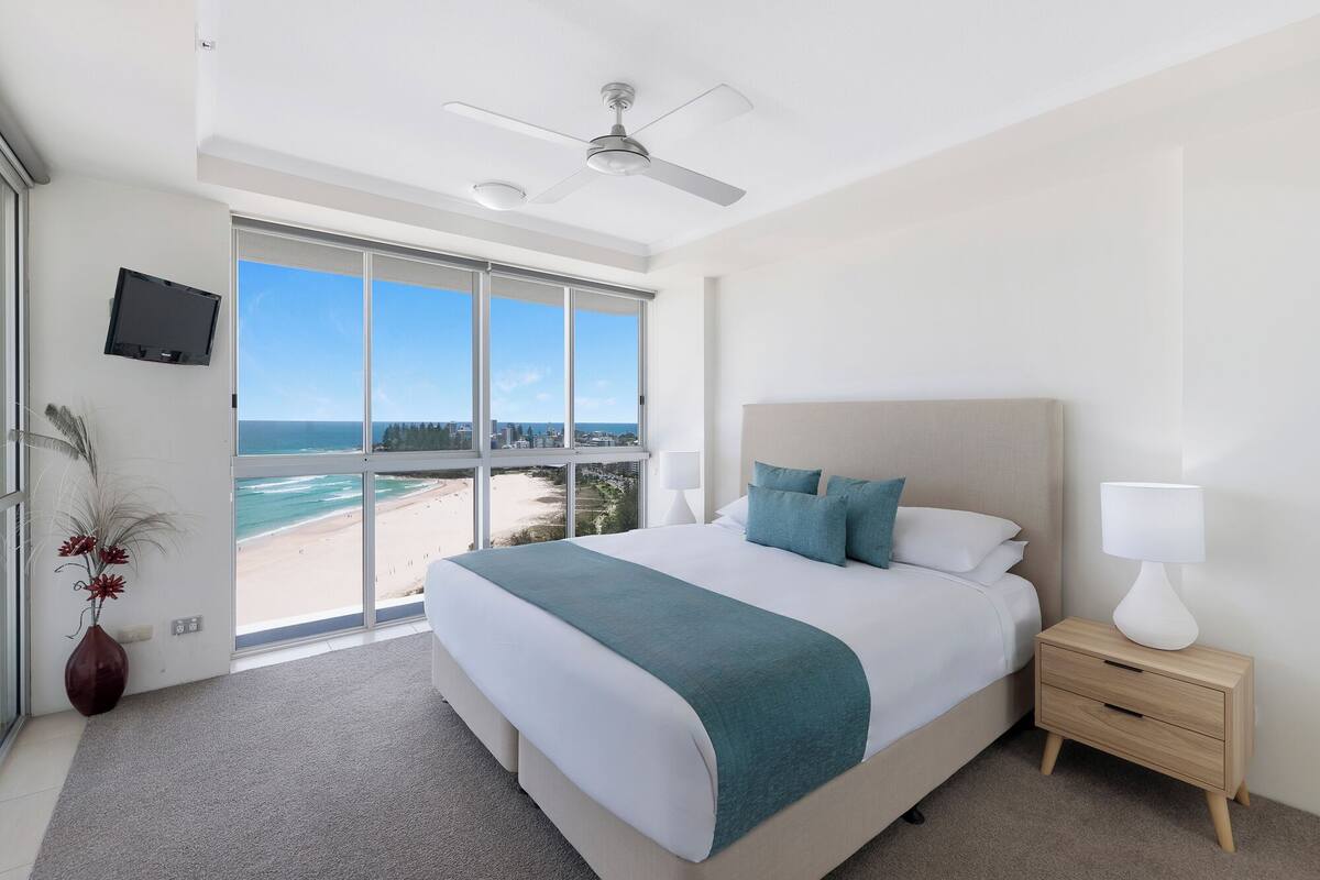 Une chambre d’hôtel dotée d’un grand lit, d’un ventilateur de plafond, d’une table de chevet avec une lampe, et offrant une vue sur la plage et l’océan grâce à de grandes fenêtres.