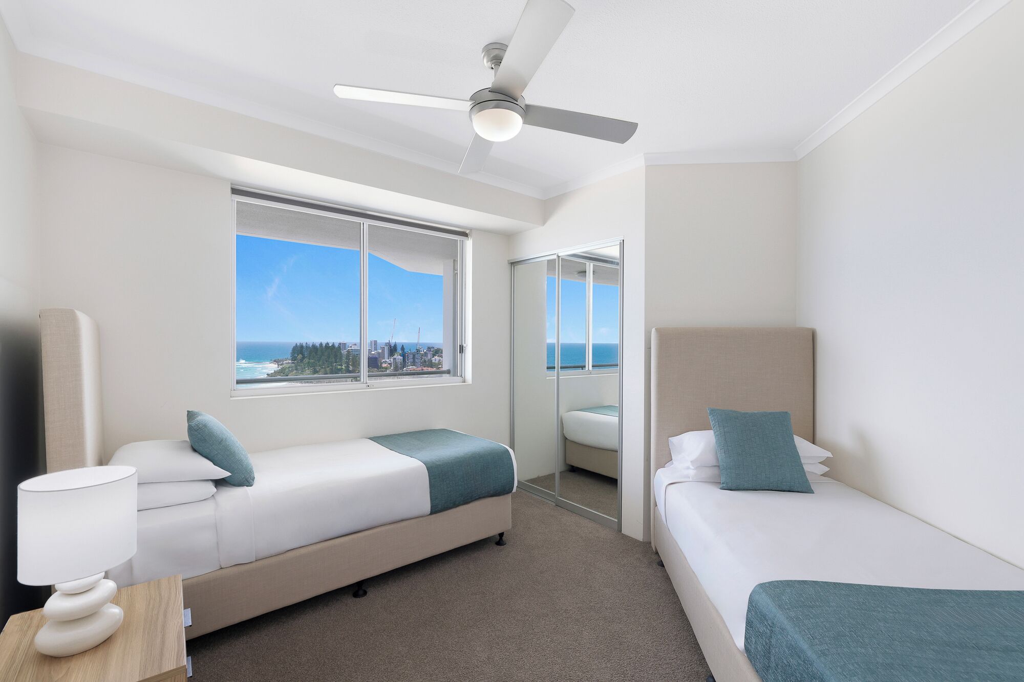 A hotel room with two beds, a ceiling fan, a large window with a view of the ocean, and a bathroom with a mirror.