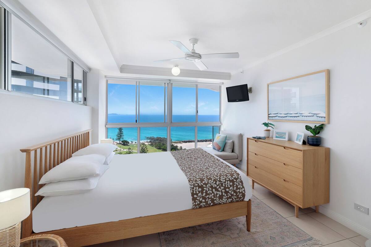 Une chambre dotée d’une grande fenêtre donnant sur la mer, d’un lit avec une literie blanche, d’une commode en bois et d’un ventilateur de plafond.