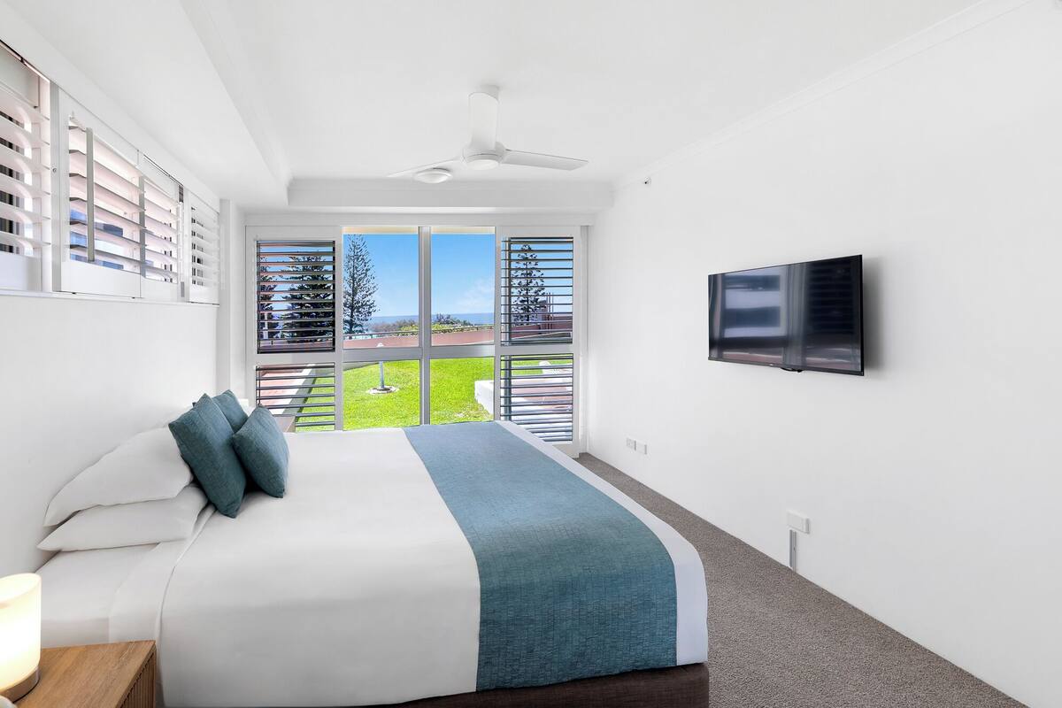 Une chambre d’hôtel moderne dotée d’un grand lit, d’un téléviseur fixé au mur et d’un balcon offrant une vue sur la ville.