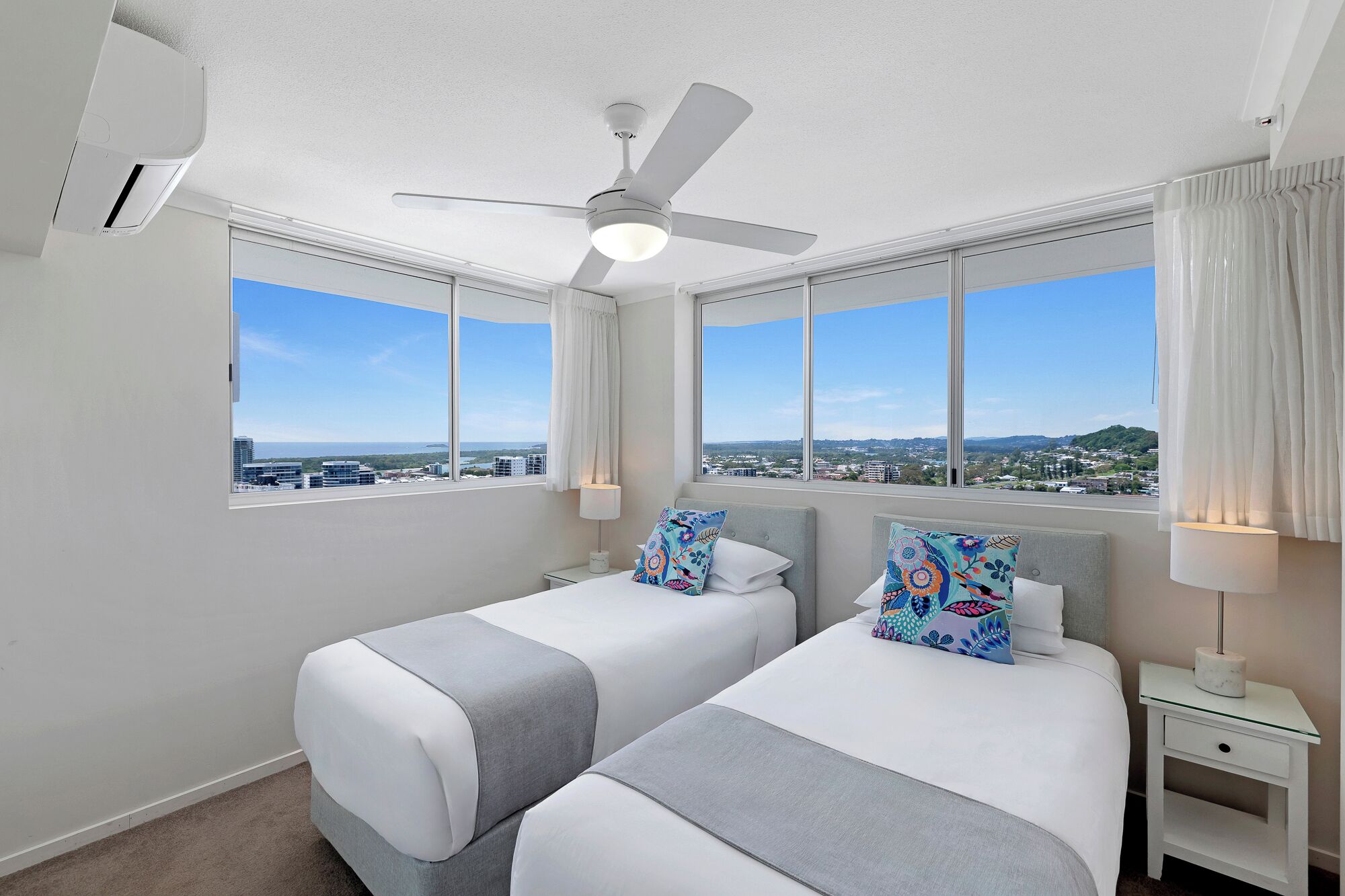 A hotel room with two beds, a ceiling fan, large windows with city views, and a nightstand with a lamp.