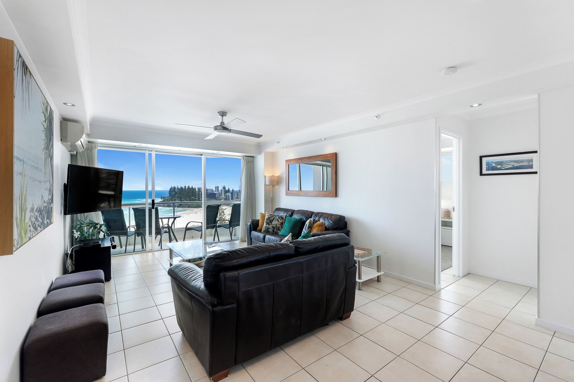 A modern living room with a black leather sofa, a flat-screen TV, and a balcony with a view of the beach.