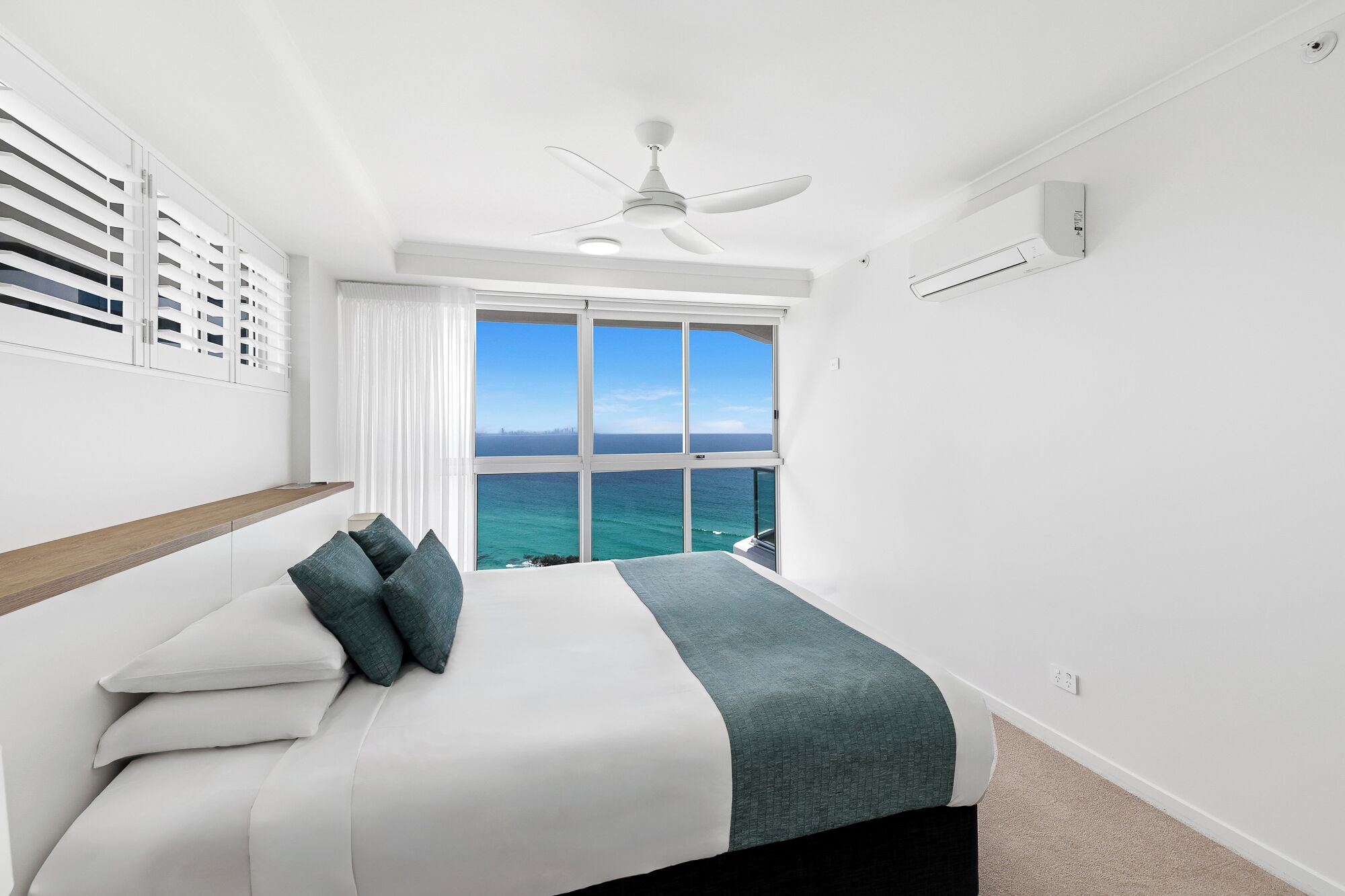 A bedroom with a bed, a ceiling fan, a window with a view of the ocean, and a white wall.