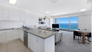 A modern kitchen with white cabinets, a built-in dishwasher, and a large island. There is a dining area with a table and chairs, and a large window offering a view of the ocean. - ULTIQA Points North (Queensland)
