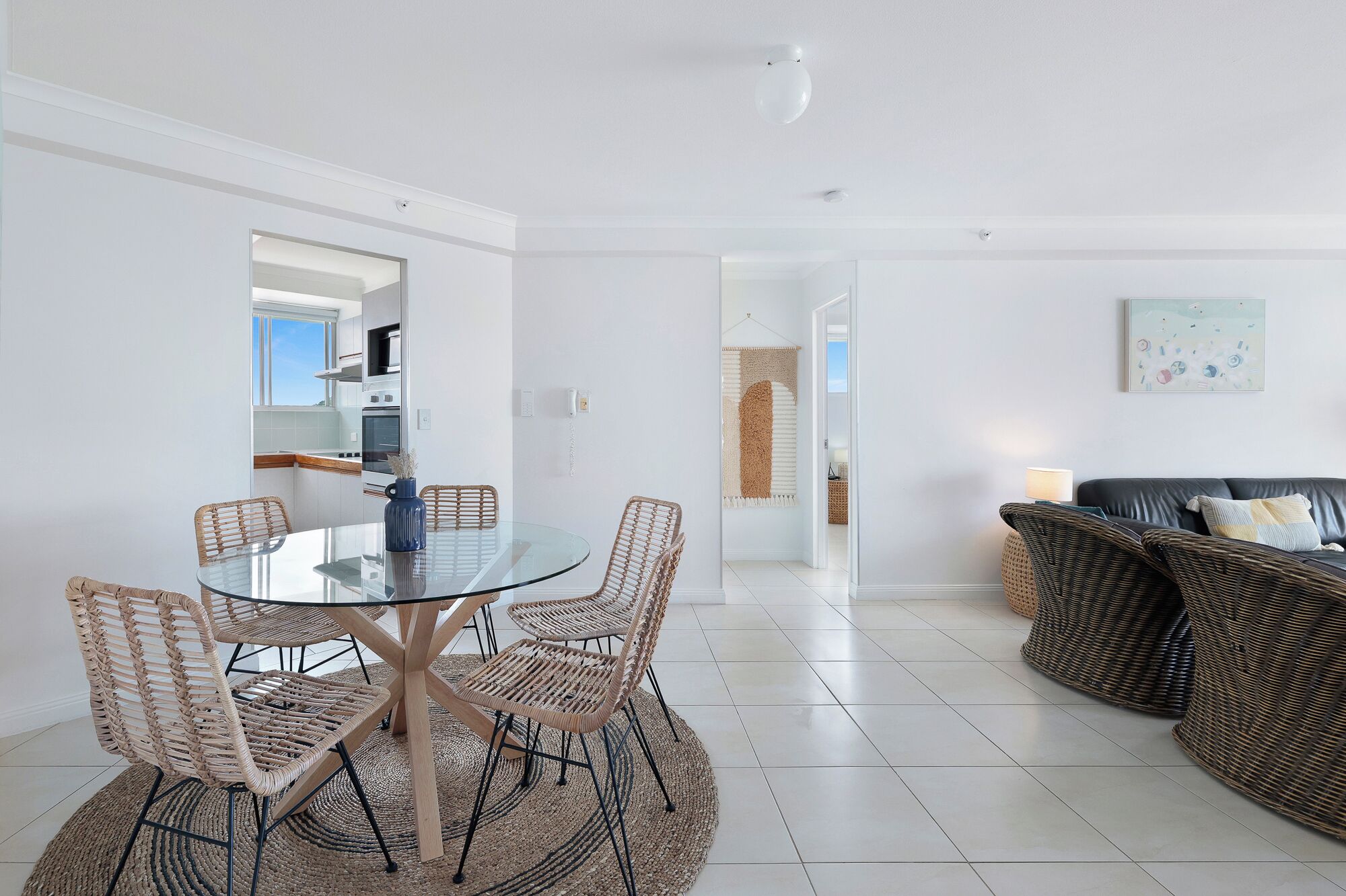 A modern living room with a glass dining table surrounded by wicker chairs, a black leather sofa, and a woven rug.