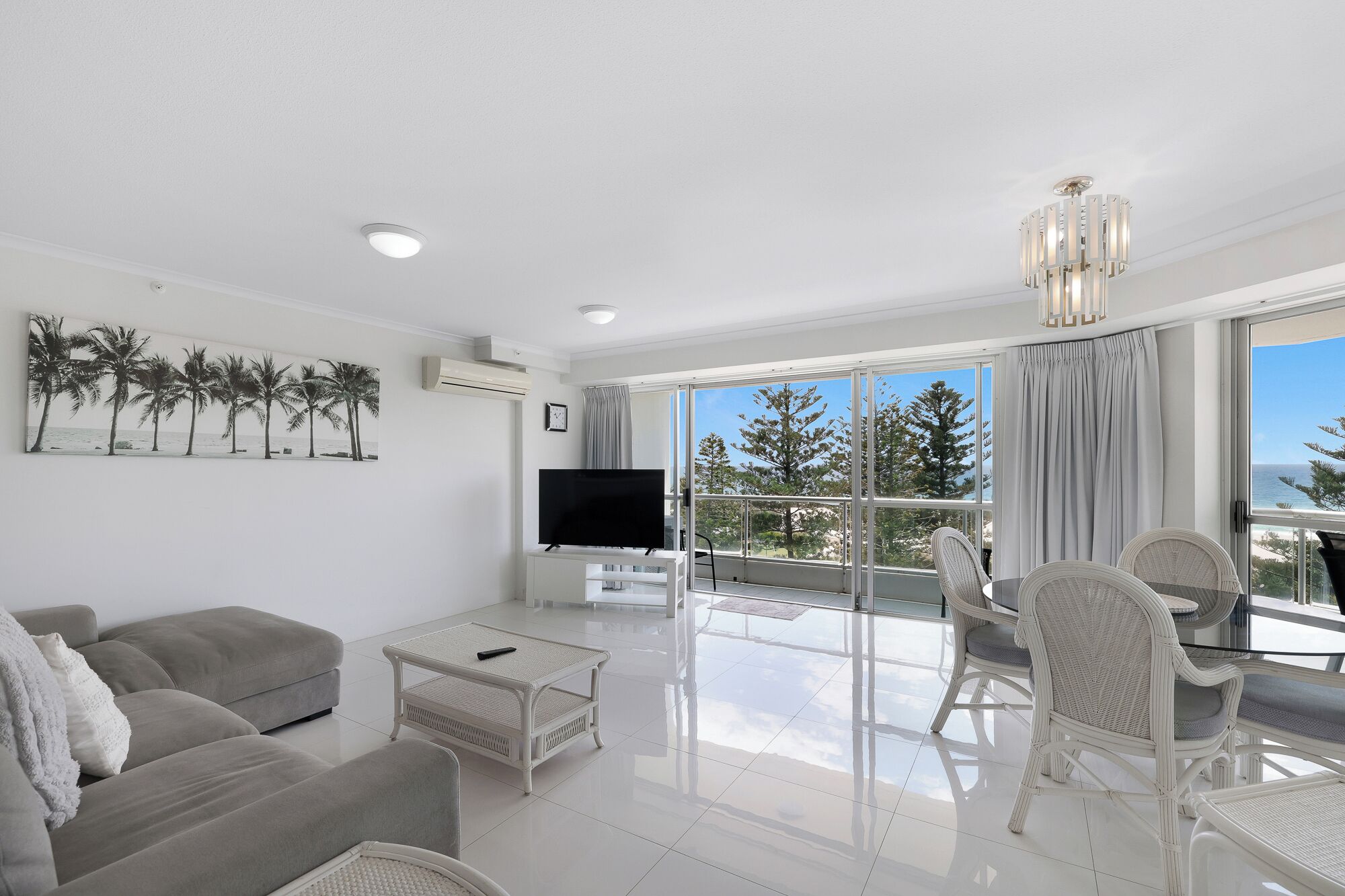 A modern living room with a flat-screen TV, a sofa, a coffee table, and a balcony with wicker chairs.