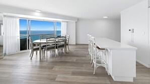 A modern kitchen with a large island, a dining area with a view of the sea, and a balcony with white curtains. - ULTIQA Points North (Queensland)