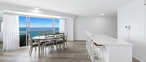 A modern kitchen with a large island, a dining area with a view of the sea, and a balcony with white curtains.