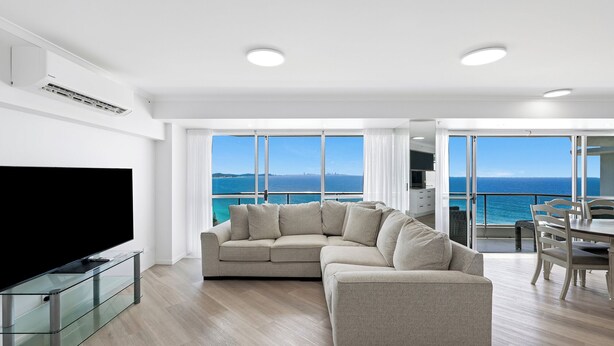 A modern living room with a sectional sofa, a glass coffee table, and a flat-screen TV. The room features large windows with a view of the sea and a balcony with outdoor seating. - ULTIQA Points North (Queensland)