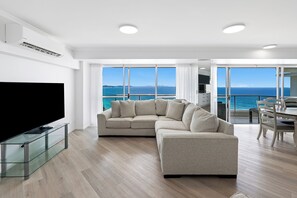 Uma sala de estar moderna com um sofá seccional, uma mesa de centro de vidro e uma TV de tela plana. O espaço possui janelas amplas com vista para o mar e uma varanda com área de estar ao ar livre.