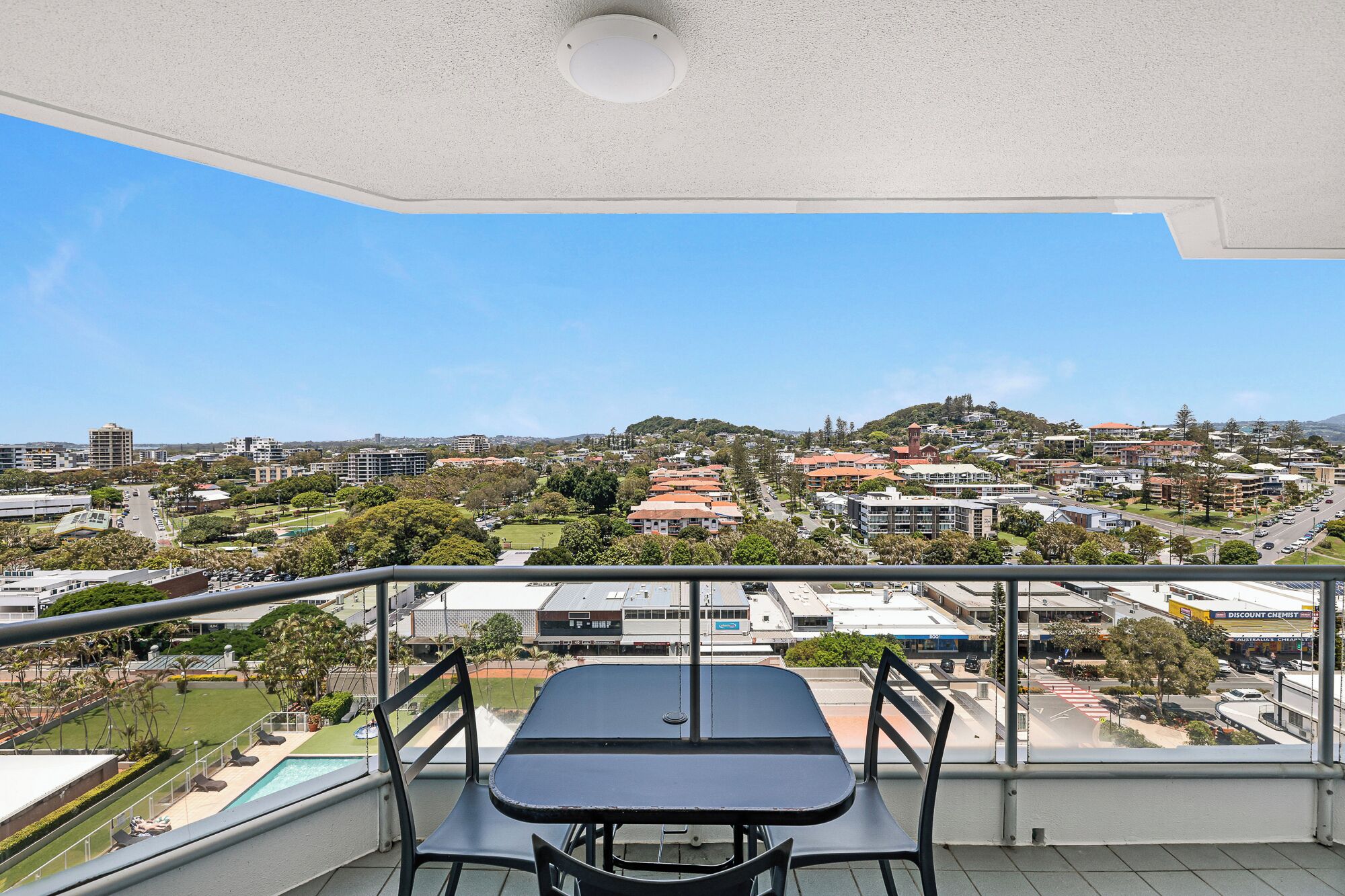Un balcon avec une table et des chaises, donnant sur un quartier résidentiel avec des maisons, une église et une colline au loin.