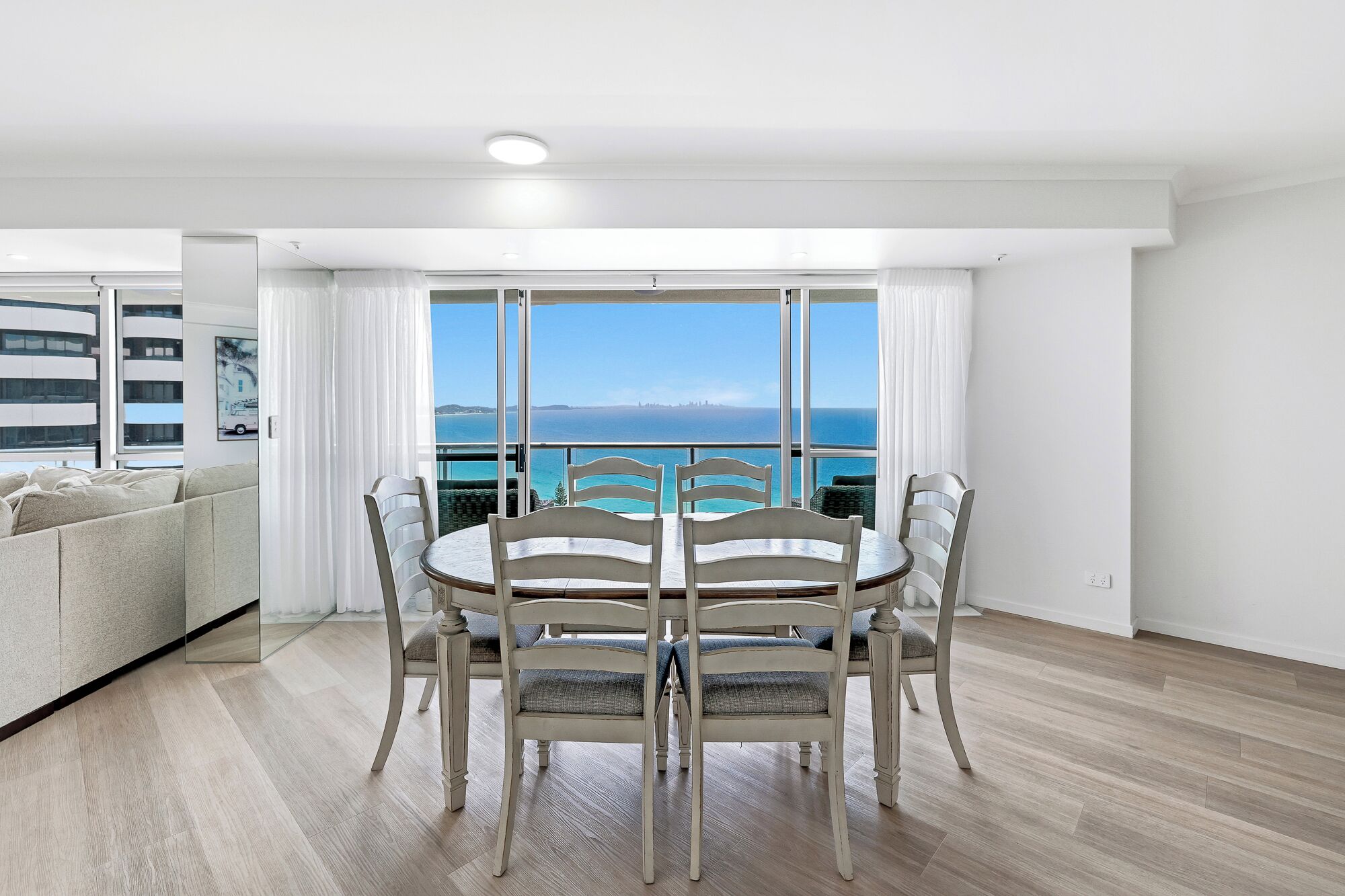 A dining area with a table and chairs, large windows with a view of the sea, and a light-colored floor.