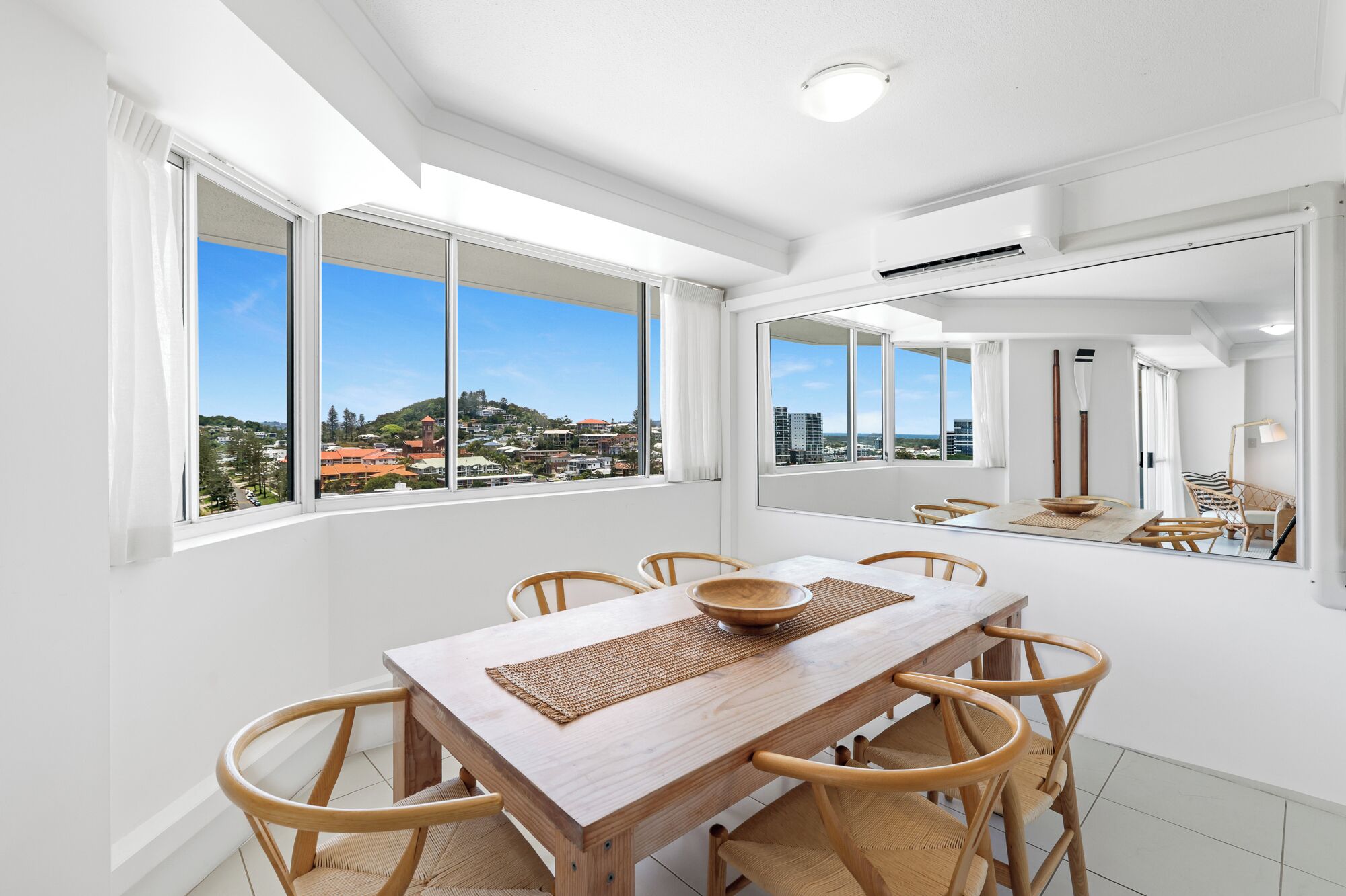 A modern dining area with a wooden table and chairs, large windows with a city view, and a ceiling light.