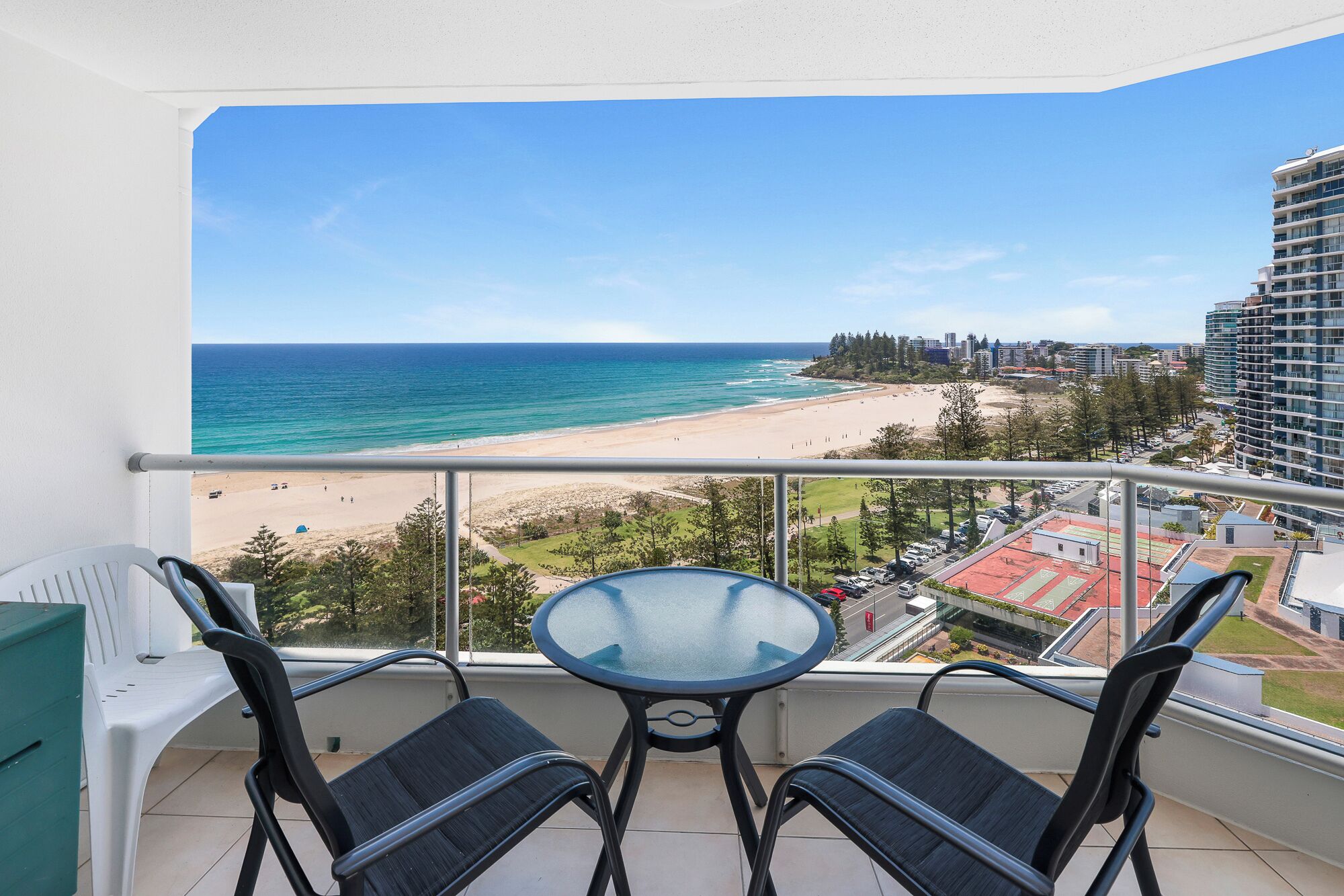 A balcony with a glass table and two chairs, overlooking a beach and cityscape.