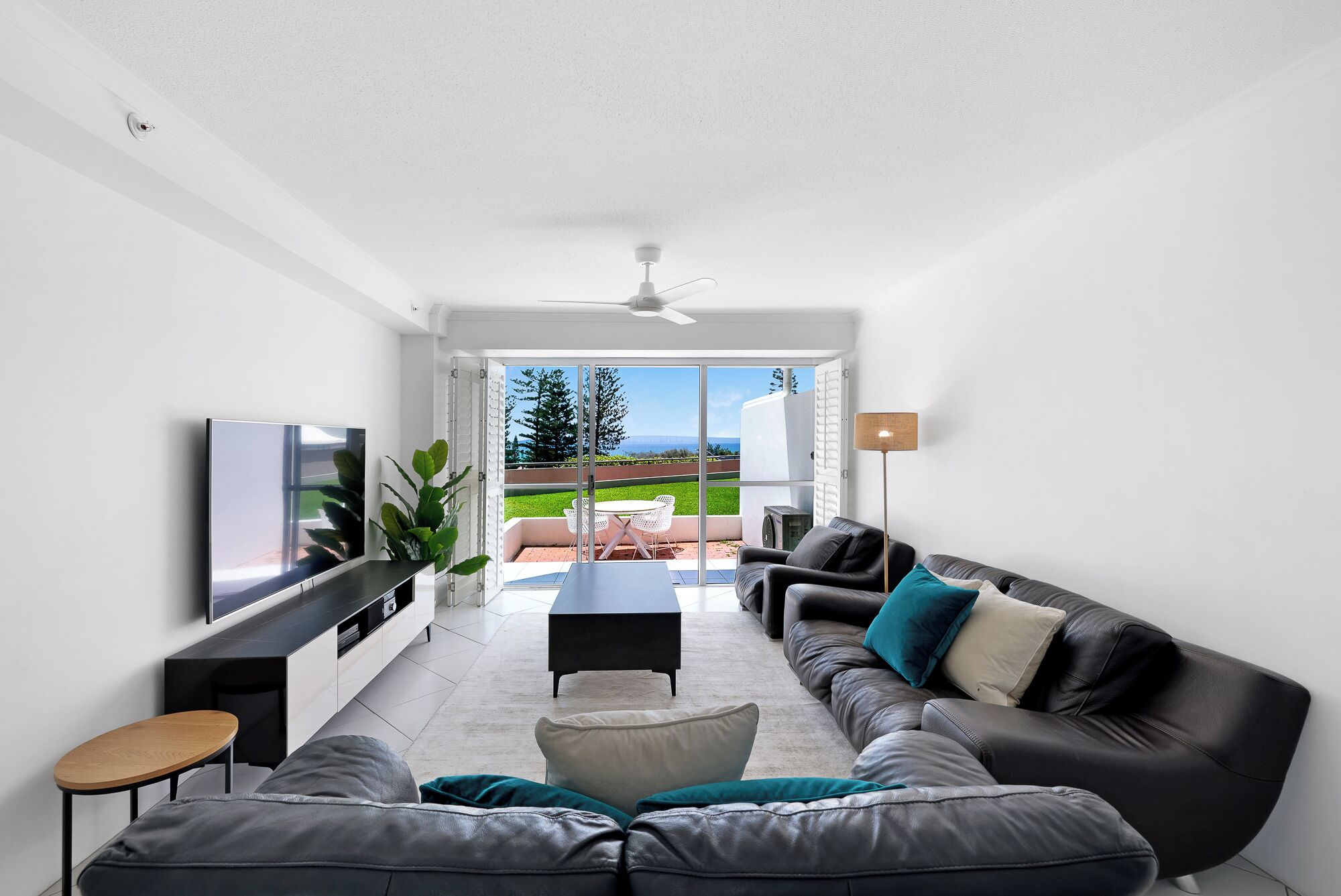 A modern living room with a sofa, coffee table, and TV. There is a sliding glass door leading to a patio with a view of the outdoors.