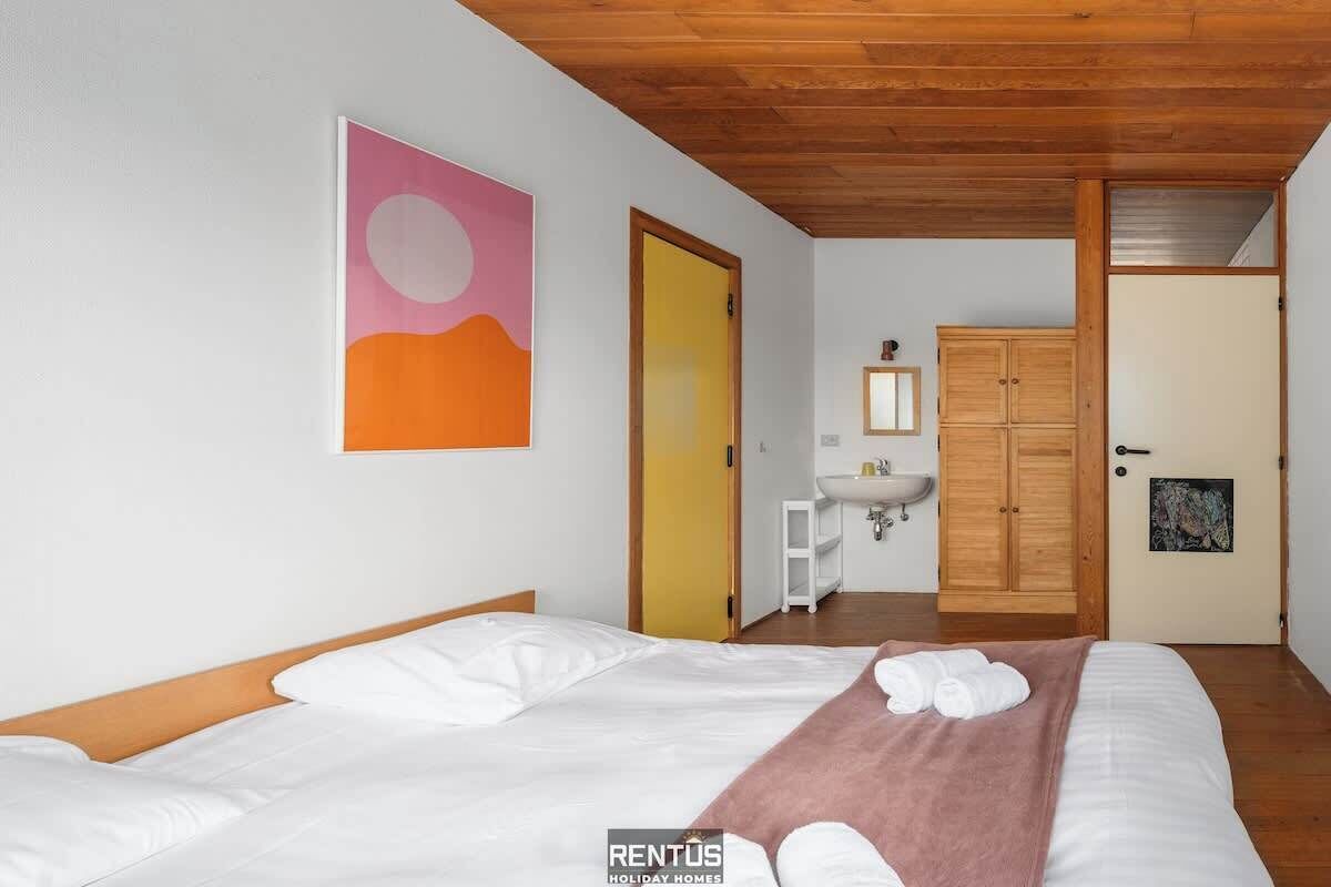 A bedroom with a wooden ceiling, a bed with white bedding, a pink blanket, and a painting of a sunset.