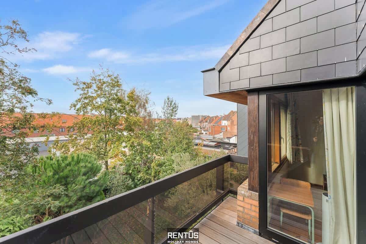 A modern balcony with a glass door, wooden deck, and a view of surrounding buildings and trees.