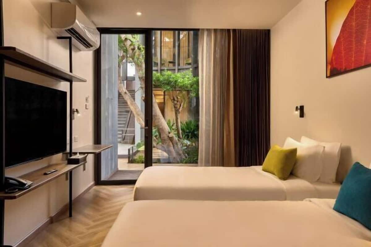 A hotel room with a TV, a bed with pillows, and a sliding glass door leading to a balcony with a view of a staircase and greenery.