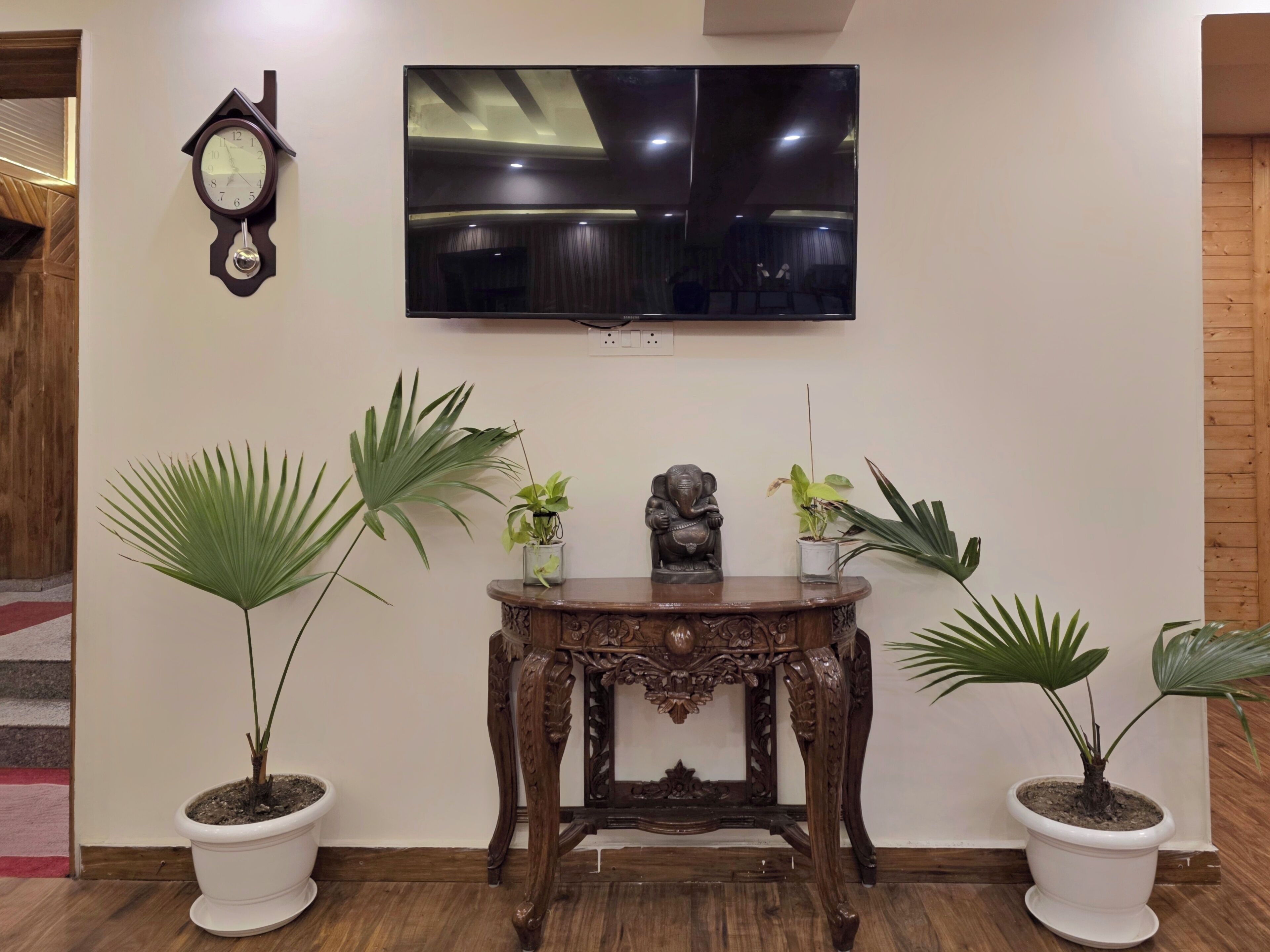 A room with a wooden floor, a TV mounted on the wall, a clock, and two potted plants on either side of a wooden table.