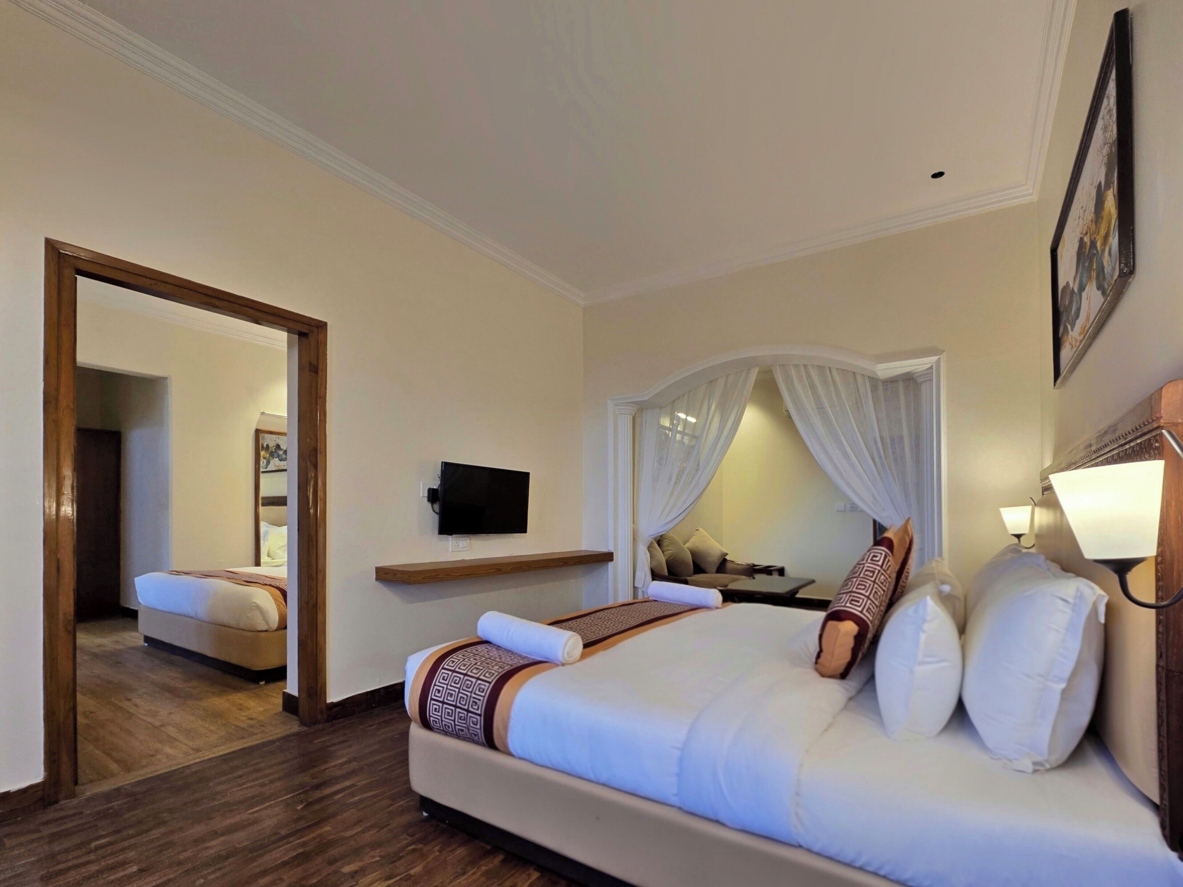 A hotel room with a large bed, a TV mounted on the wall, a bedside table with a lamp, and a mirror.