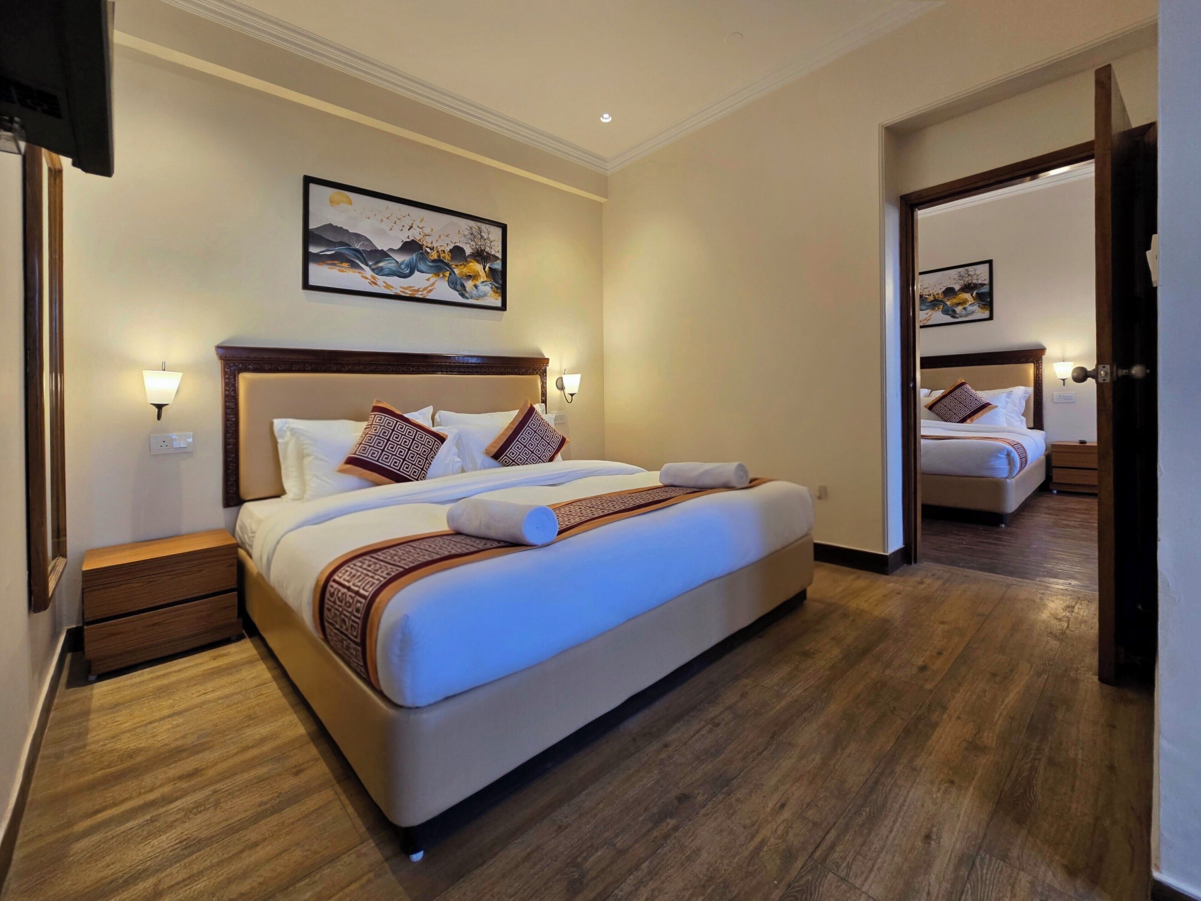 Una camera d'hotel con un letto grande, una testiera in legno e un quadro sopra. C'è un comodino con una lampada e un cassetto in legno. La stanza ha un pavimento in legno e una porta che conduce a un'altra stanza.