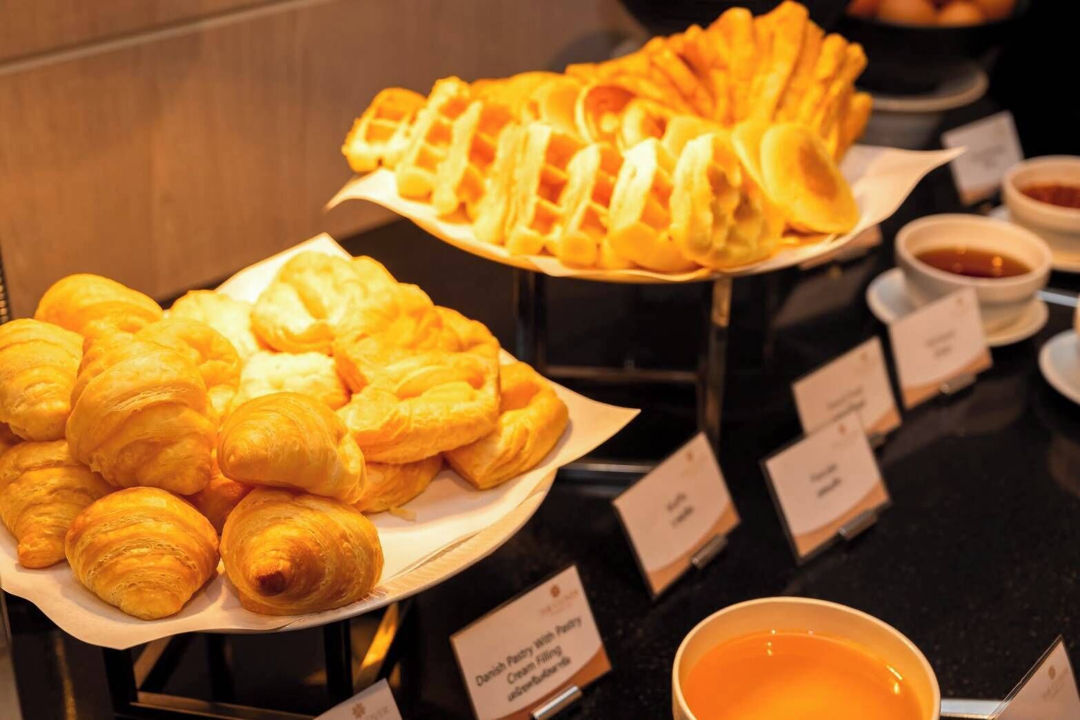 A display of pastries including croissants and waffles, with small bowls of sauces and a sign indicating Danish pastry with cream filling.