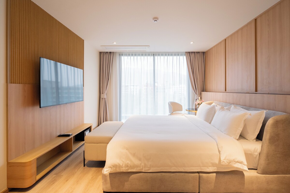 A modern hotel room with a large bed, a TV mounted on the wall, a wooden panel design, and a large window with curtains.