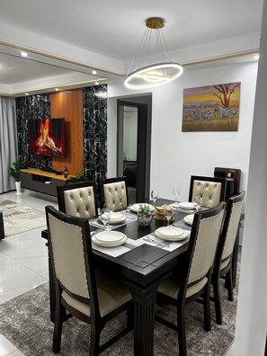 A dining area with a black table set for six, surrounded by upholstered chairs. A flat-screen TV is mounted on the wall above the fireplace, and a painting of zebras is visible on the wall.