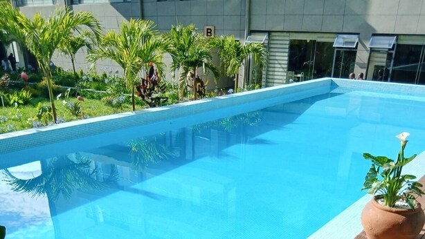 A swimming pool with a wooden deck, surrounded by greenery and a building in the background.