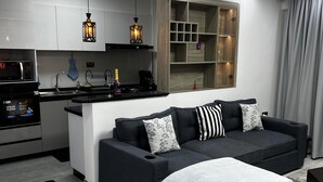 A modern living room with a dark grey sofa, a white rug, and a kitchen area with a microwave, stove, and pendant lights.