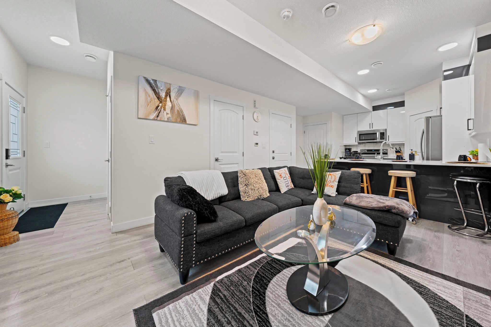 A modern living room with a grey sofa, a glass coffee table, and a kitchen in the background.