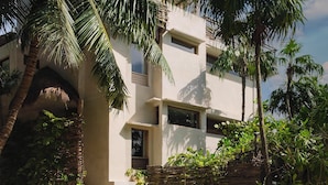 Un bâtiment moderne à deux étages, doté d’un toit plat et entouré d’une végétation luxuriante, notamment de palmiers et d’autres plantes tropicales.