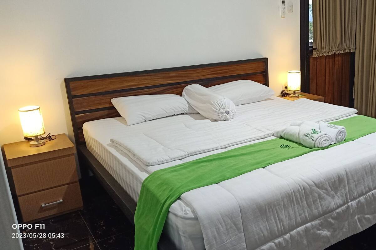 Ein ordentlich bezogenes Bett mit weißen Leinen und einer grünen Tagesdecke, ein Holz-Kopfteil, Nachttischlampen, ein kleiner Holzschemel und ein Marmorboden.